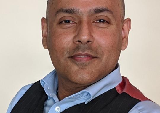 Headshot of DR UK CEO Kamran Mallick, he is wearing a light blue shirt, with a black suit vest.
