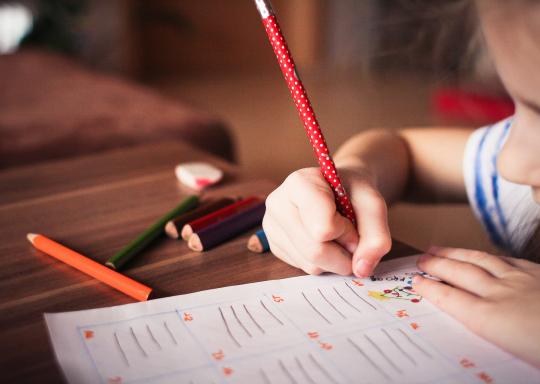 image shows a young girl leaning on a wooden desk colouring in a school book.