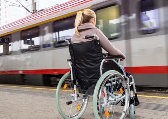 image shows the back of a wheelchair user. She is facing a train as it arrives in front of her. She has blond hair in a ponytail.