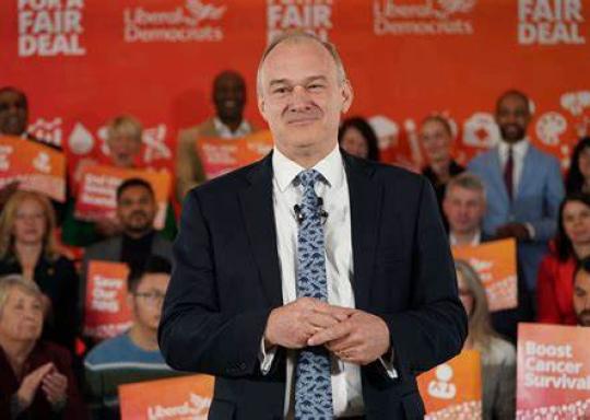 Ed Davey, wearing a blue shirt, blue tie and white shirt, is standing in front of a crowd of Lib Dem supporters. 