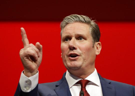 Keir Starmer wearing a dark blue suit, white shirt and red tie is in front of an all red background.