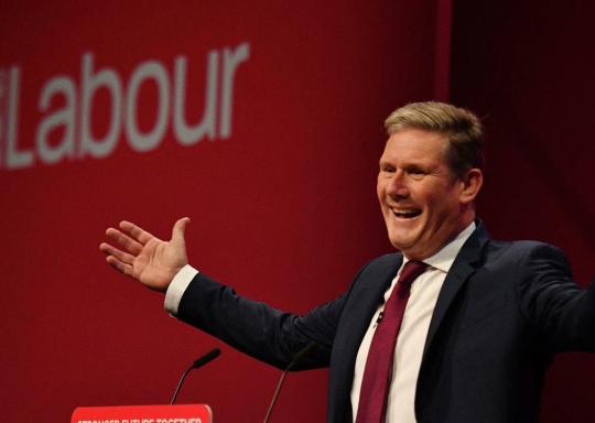 Keir Starmer stands hand raised and smiling in front of a red background with the words 