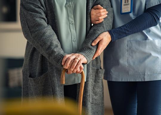 A nurse is supporting an elderly person who wears a grey cardigan and who is holding a walking stick