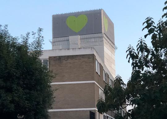Grenfell Tower is pictured, it is a tall modernist tower block with a smaller georgian house in the foreground, Grenfell is covered in a green covering with yellow heart in the middle.